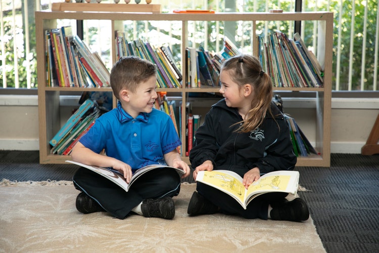 2 students reading a book in the library and smiling at each other